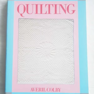以下が含まれることがあります： Averil Colby著のハードカバー本「Quilting」。表紙はグレーの角を持つ水色の縁取り、ピンクの中央パネル、そして複雑な花柄とダイヤモンド模様がステッチされた白いキルトの写真で構成されています。