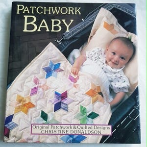 May include: A hardback book titled Patchwork Baby by Christine Donaldson featuring a cover image of a baby lying in a dark pram under a cream-coloured quilt decorated with colourful, multi-pointed star-shaped patchwork patterns.