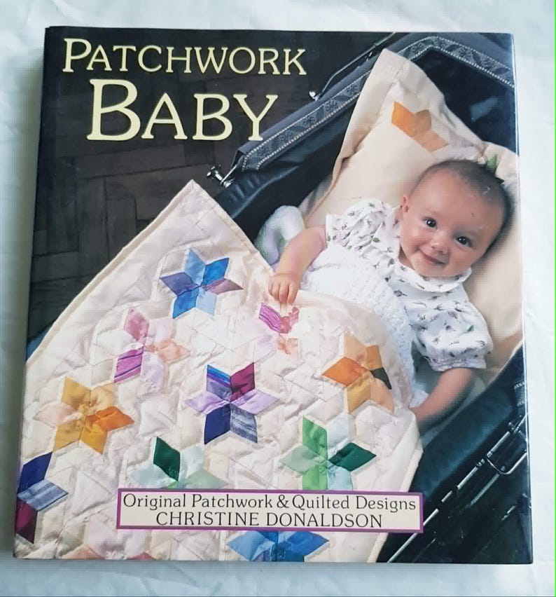 Może przedstawiać: Książka w twardej oprawie zatytułowana Patchwork Baby autorstwa Christine Donaldson, z okładką przedstawiającą dziecko w ciemnym w&oacute;zku pod kremową kołdrą ozdobioną kolorowymi, wieloramiennymi wzorami patchworkowymi w kształcie gwiazd.