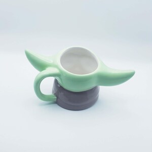 May include: A ceramic mug shaped like a character with large pointed ears, a light green upper section, and a dark gray base. The mug features a curved handle on the side and a wide, circular opening at the top, designed with a smooth, glossy finish.