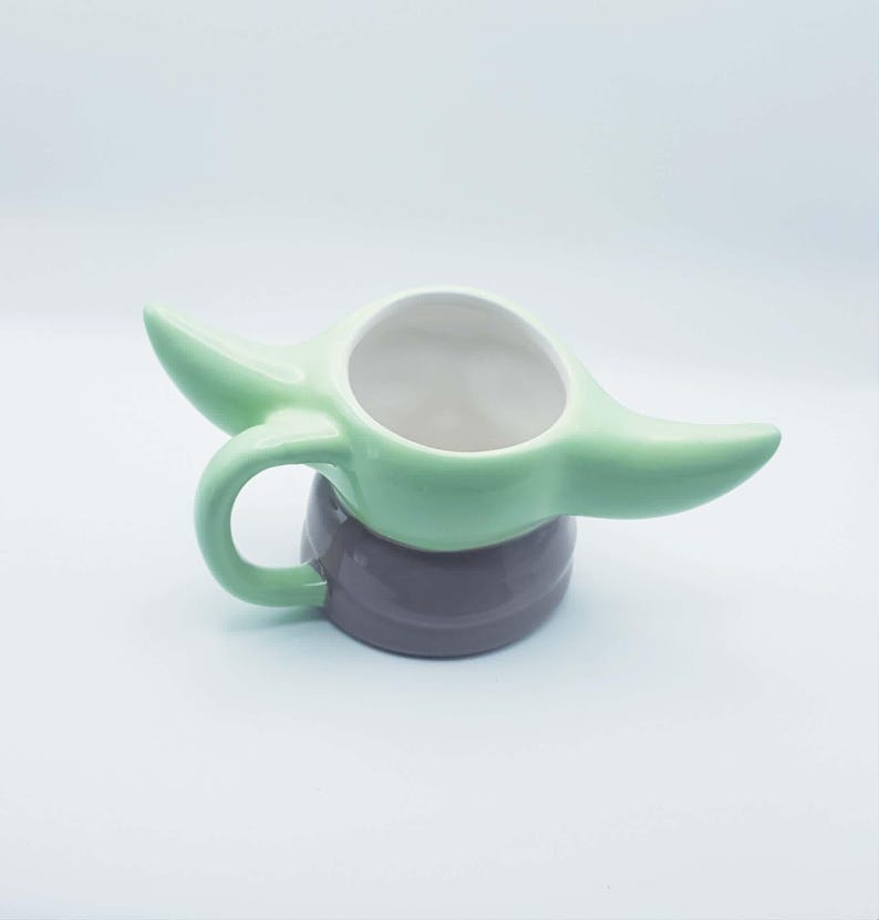 May include: A ceramic mug shaped like a character with large pointed ears, a light green upper section, and a dark gray base. The mug features a curved handle on the side and a wide, circular opening at the top, designed with a smooth, glossy finish.