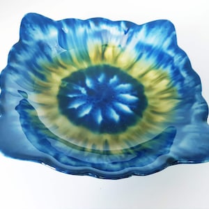 May include: A square glass bowl with wavy, scalloped edges. The bowl features a tie-dye pattern with a deep blue center, transitioning into a soft yellow ring, and ending with a dark blue border. The glass has a smooth, glossy finish.