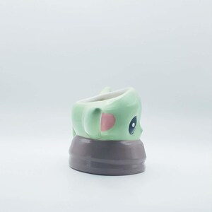 May include: A ceramic mug shaped like a small green creature with large dark eyes and pink inner ears. The creature is wearing a brown collar or robe base. The mug has a smooth, glossy finish and a white interior, viewed from a side profile angle.