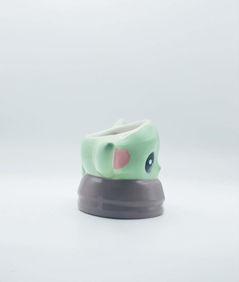 May include: A ceramic mug shaped like a small green creature with large dark eyes and pink inner ears. The creature is wearing a brown collar or robe base. The mug has a smooth, glossy finish and a white interior, viewed from a side profile angle.