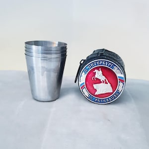 May include: A set of four small, silver-colored metal shot glasses next to a black leather case with a circular emblem. The emblem features a red center with a white horse and rider, surrounded by text and a blue and white border.