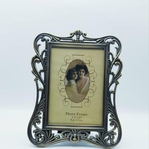 May include: Ornate bronze-colored photo frame. The frame has an intricate, scrolling design around the edges. The frame holds a sepia-toned photograph of two people. The text "Photo Frame 3.5"x5" 9cm x 13cm" is visible.