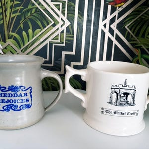 May include: Two ceramic mugs. One is light gray with a blue label that reads "Cheddar Rejoices". The other is white with a black illustration of a building and the text "The Market Cross".