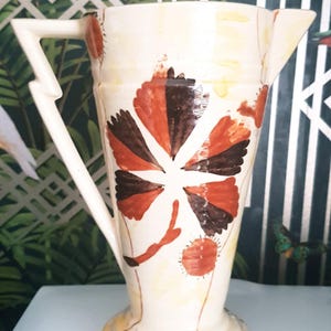 May include: Cream-colored ceramic pitcher with a unique lightning bolt handle and a spout. The pitcher is decorated with a stylized floral design in shades of brown, orange, and red. The pitcher has a bulbous base and a tall, cylindrical body.