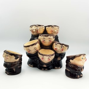 May include: A ceramic set of seven monk-shaped shot glasses. The glasses are brown and beige with smiling faces and black robes. The set includes a larger base with six glasses and one separate glass.