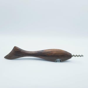 May include: A dark brown wooden corkscrew shaped like a fish, featuring a carved tail and a metal spiral worm extending from the mouth. A small metal bottle opener is integrated into the underside of the fish-shaped body, providing a dual-function tool.