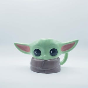 May include: A ceramic mug shaped like a small green creature with large black eyes, pointed ears with pink inner details, and a brown robe-like base. The mug features a handle on the right side and a smooth, glossy finish throughout the entire piece.