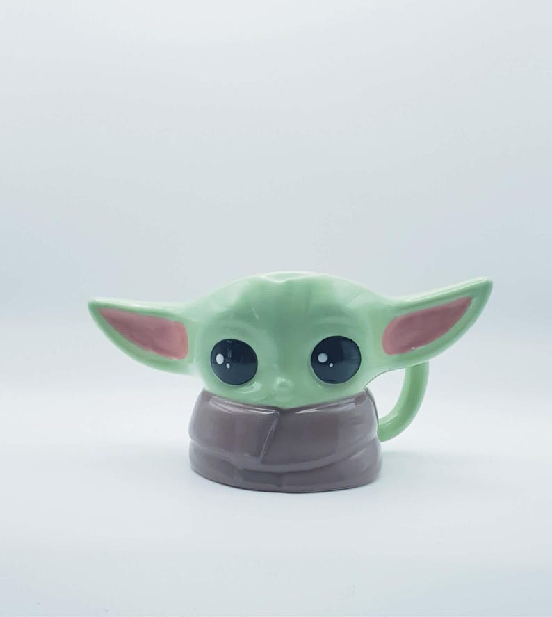 May include: A ceramic mug shaped like a small green creature with large black eyes, pointed ears with pink inner details, and a brown robe-like base. The mug features a handle on the right side and a smooth, glossy finish throughout the entire piece.