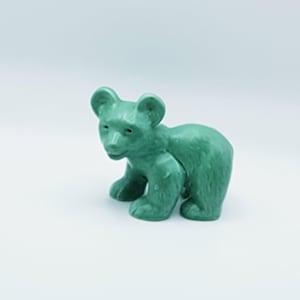 Könnte beinhalten: Eine kleine, grüne Bärenfigur. Der Bär wird in einer Laufhaltung dargestellt, mit den Vorderpfoten nach vorne. Die Figur hat eine strukturierte Oberfläche und abgerundete Merkmale, einschließlich kleiner Ohren und einer kurzen Schnauze. Der Bär hat eine einfarbige, opake grüne Farbe.