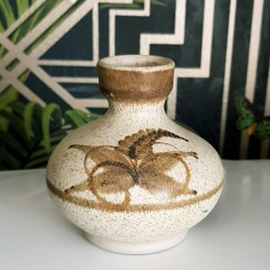 May include: A small, speckled ceramic vase with a rounded body and a narrow neck. The vase features brown floral designs and a brown rim. The background includes a green and white geometric pattern and a butterfly.