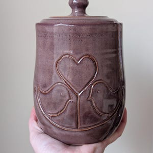 May include: A mauve-colored ceramic canister with a matching lid and a knob handle. The exterior features a debossed design of two birds facing each other with a heart shape positioned between them, all outlined in a lighter shade of mauve.