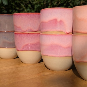 Pode incluir: Uma coleção de sete copos de cerâmica dispostos em filas. Cada copo apresenta uma base bege lisa sem verniz e uma secção superior brilhante mergulhada em vários tons de rosa, desde pastel suave a magenta profundo, com padrões de gotas visíveis.