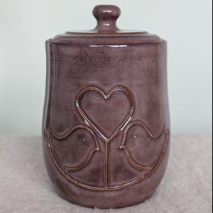 Op de afbeelding: Een keramische pot met een deksel, met een hart- en twee vogels ontwerp. De pot is diep paars van kleur met een glanzende afwerking.