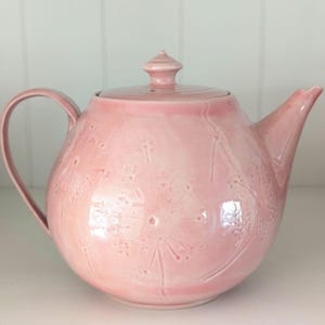 Op de afbeelding: Een lichtroze keramische theepot met een glanzende afwerking en een ronde vorm. Het oppervlak is voorzien van reliëfpatronen van paardenbloempluisjes. De theepot heeft een bijpassend deksel met een knop, een gebogen tuit en een zijhandvat.