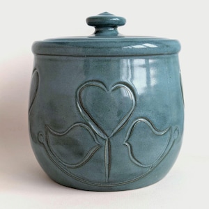 Può includere: Un barattolo in ceramica turchese con coperchio, con un motivo a cuore e uccello stilizzato in rilievo. Il barattolo ha una forma arrotondata e una finitura lucida. Il coperchio ha un piccolo pomello. Un contenitore decorativo per la cucina.