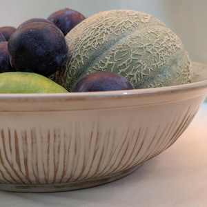 Op de afbeelding: Een beige keramische kom gevuld met vers fruit. De kom bevat pruimen, een cantaloupe, een groene peer en een sinaasappel. De kom heeft een textuur, geribbeld ontwerp aan de buitenkant.