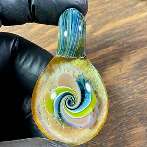 May include: A glass pendant with a swirl design in shades of blue, green, pink, and yellow. The pendant is surrounded by a textured, golden-brown background.