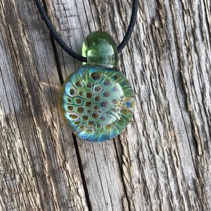 Puede incluir: Un colgante de vidrio verde y azul con una superficie texturizada, colgando de un cordón negro.