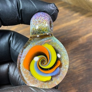 May include: A glass pendant with a swirling pattern of orange, yellow, blue, and black. The pendant has a iridescent finish and is set against a dark background.
