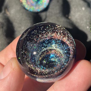 Puede incluir: Una canica de vidrio esférica oscura con un diseño de galaxia arremolinada. La canica presenta un fondo azul oscuro y negro con purpurina incrustada en tonos plateados, azules y rosas. Otra canica con un diseño de arcoíris es visible en el fondo.
