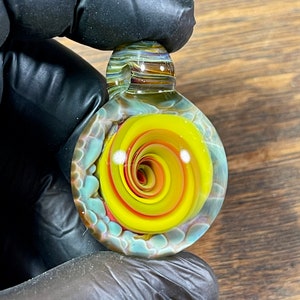May include: A glass pendant with a yellow, orange, and red spiral design inside. The pendant is clear and has a blue and white swirl pattern around the outside.