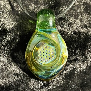 Puede incluir: Un colgante de vidrio verde con un patrón de remolino de amarillo, azul y blanco. El colgante tiene una pequeña abertura redonda en el centro con un patrón de círculos blancos y azules.