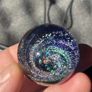 Puede incluir: Una esfera de vidrio transparente con un diseño de galaxia en espiral. La esfera presenta tonos de azul, verde y purpurina iridiscente, creando un efecto de cosmos en miniatura. La esfera se sostiene en una mano.