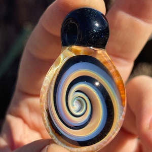 May include: A glass pendant with a black and white spiral design. The spiral is surrounded by a ring of brown glass. The pendant has a black glass top.