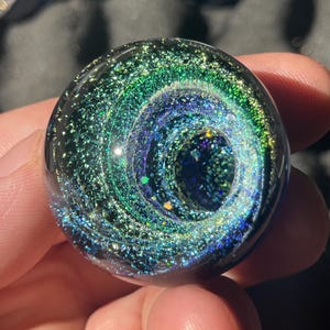 Puede incluir: Una canica de vidrio redonda y transparente con un diseño de galaxia arremolinada. La canica presenta tonos de verde, azul y purpurina plateada, creando un efecto cósmico. El diseño está encerrado en vidrio transparente.