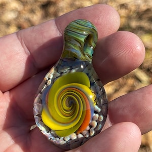 Puede incluir: Un colgante de vidrio con un remolino de vidrio amarillo, naranja y azul en su interior. El colgante es transparente y tiene un remolino verde, azul y marrón en la parte superior.