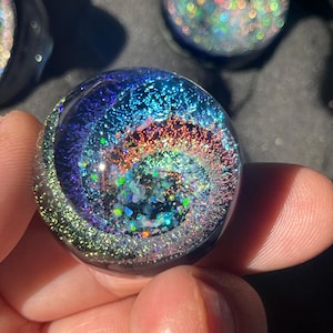 Puede incluir: Una canica de vidrio redonda y transparente con un diseño de galaxia en espiral. El interior presenta una mezcla de purpurina azul, morada, verde y roja, creando un efecto cósmico. La canica se sostiene en una mano, con otras canicas similares en el fondo.
