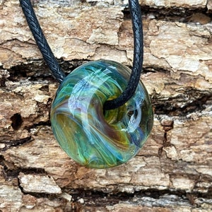 Peut inclure: Un pendentif rond en verre vert, bleu et brun avec un motif en spirale suspendu à un cordon noir. Le pendentif est rond et a un trou au centre.