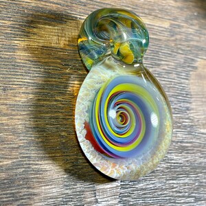 May include: A glass pendant with a swirling pattern of yellow, red, blue, and white. The pendant has a clear glass top and a green and brown glass bottom.