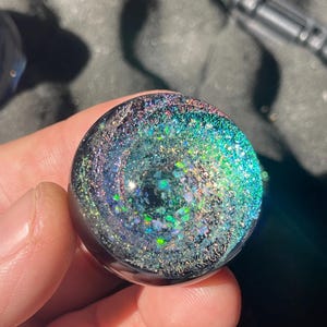 Puede incluir: Un objeto redondo de vidrio negro con un diseño de galaxia en espiral. El diseño presenta purpurina iridiscente en tonos azules, verdes y plateados. El objeto se sostiene en una mano.
