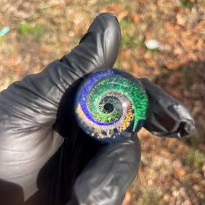 Puede incluir: Una canica de vidrio con un diseño de espiral verde, azul y dorado. La canica es transparente y tiene un acabado brillante.