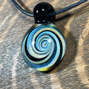 May include: A glass pendant necklace with a black cord. The pendant is a swirl of blue, yellow, and black glass.