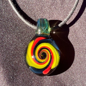 Pode incluir: Um colar com um pingente de vidro em um cordão preto. O pingente é um orbe redondo de vidro transparente com um design em espiral em vermelho, amarelo e preto.