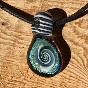 Puede incluir: Un collar con un colgante de vidrio, la parte superior es de rayas blancas y negras, y la parte inferior está decorada con un espiral azul y blanco. El colgante está unido a un cordón negro.