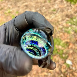 Puede incluir: Una canica de vidrio redonda con un patrón de espiral de azul, verde y dorado. La canica es iridiscente y tiene un aspecto moteado.