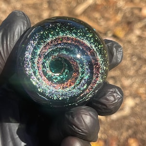 Puede incluir: Una canica de vidrio esférica oscura con un diseño de galaxia arremolinada. El interior presenta purpurina iridiscente en tonos verdes, rojos y dorados, creando un efecto cósmico. La canica se sostiene en una mano enguantada.