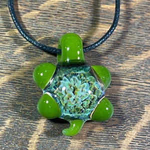 May include: A green glass turtle pendant with a detailed, multi-colored, swirling design inside. The pendant is attached to a black cord.