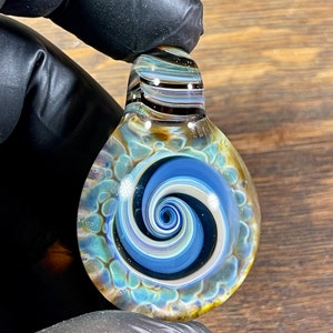 May include: A glass pendant with a swirling blue, white, and black design. The pendant has a brown and white striped top and a textured, iridescent surface.