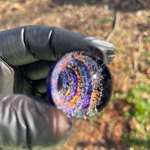 Puede incluir: Primer plano de una pequeña esfera de vidrio redonda con un patrón de remolino de purpurina púrpura, azul y dorada. La esfera se sostiene en una mano enguantada.