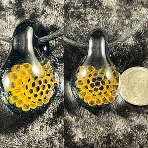 Puede incluir: Un colgante de vidrio negro con un diseño de panal de abejas en su interior. El colgante es aproximadamente del tamaño de una moneda de 25 centavos de dólar estadounidense.