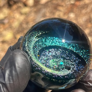 Puede incluir: Una esfera de vidrio transparente con un diseño interno en espiral de purpurina turquesa y plateada. La esfera se sostiene en una mano enguantada, con la luz del sol reflejada en la superficie. El efecto general recuerda a una galaxia.