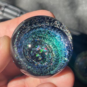 Puede incluir: Un pisapapeles esférico de vidrio transparente con un diseño de galaxia interno. El interior presenta remolinos de azules, verdes y purpurina iridiscente, creando un efecto de cosmos en miniatura. La superficie exterior es lisa y reflectante, con un fondo oscuro.